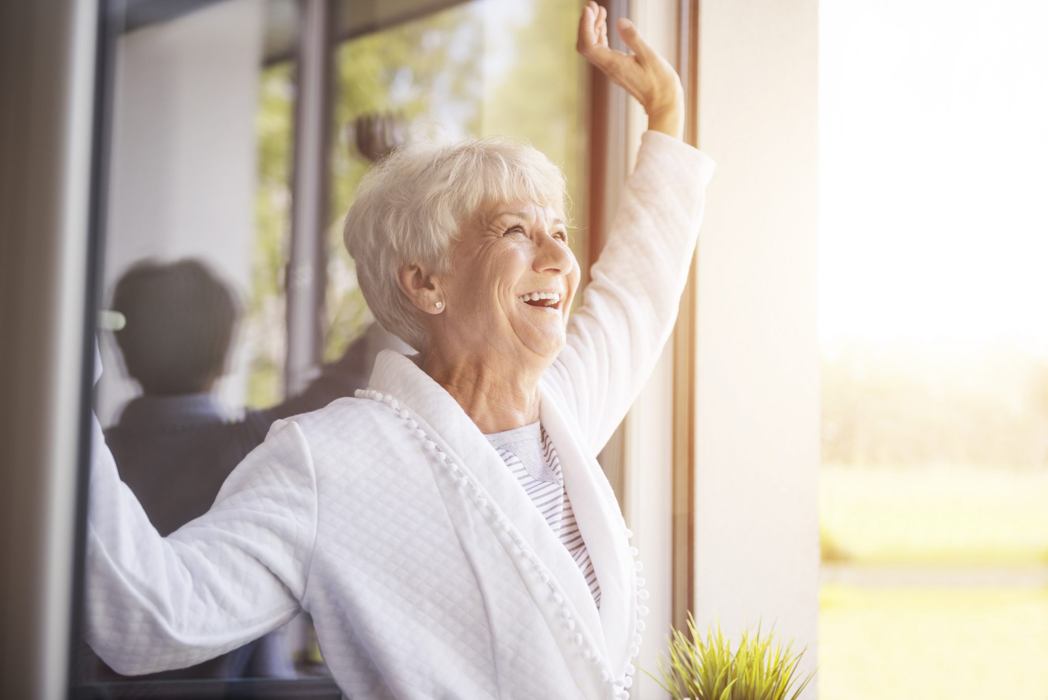 Smiling elderly woman raising arm while looking out a sunny window