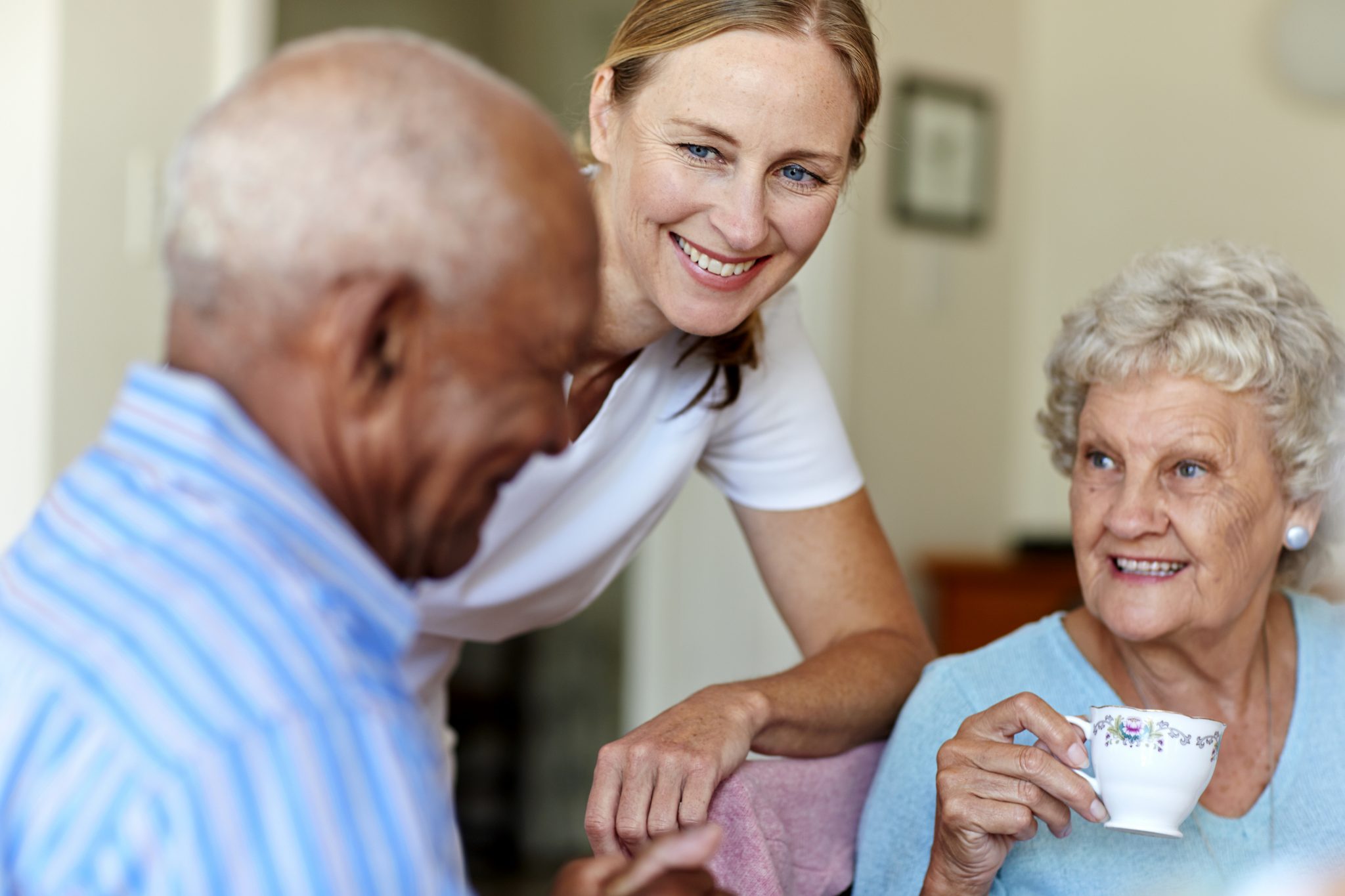 Woman smiling at two seniors having tea in a cozy living space.