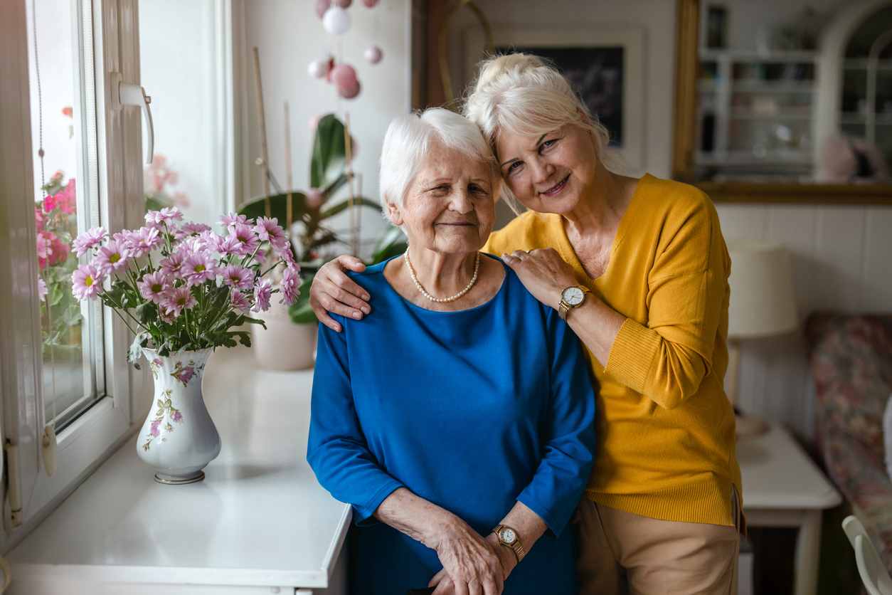 Two elderly women smiling together in a cozy living space with flowers on the table.