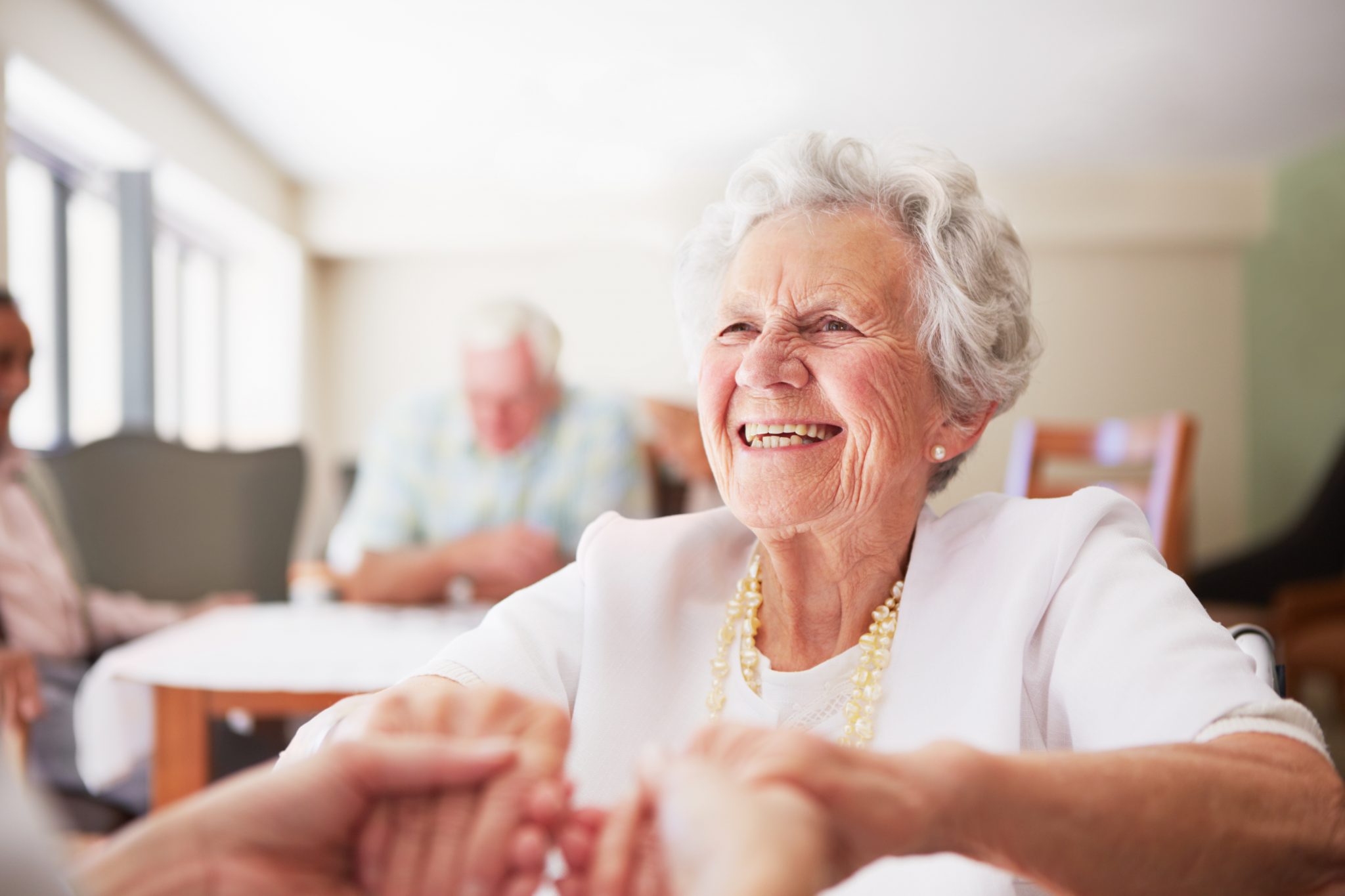 Elderly woman smiling and holding hands in a communal room of a living space.