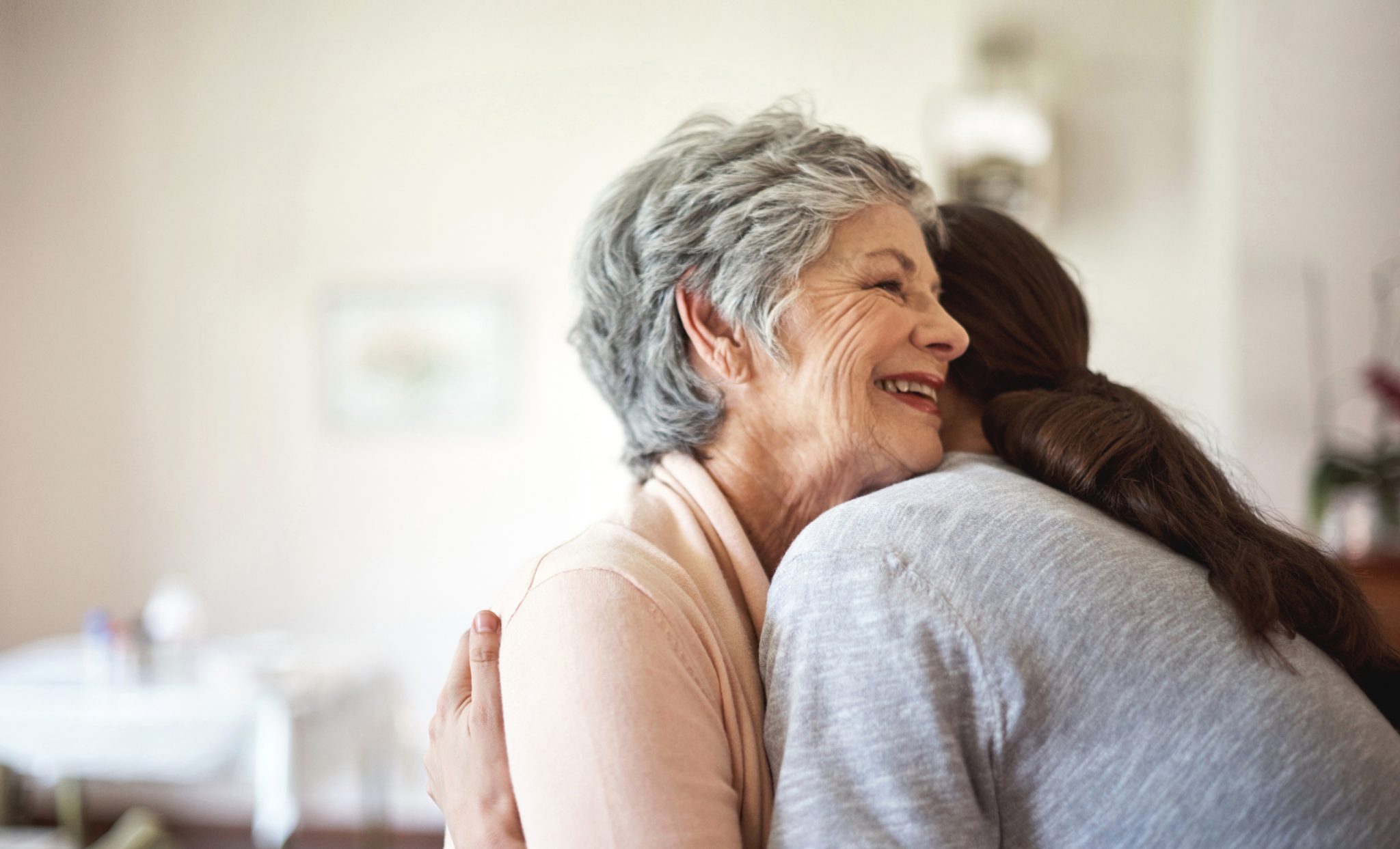 Elderly woman joyfully hugging young woman in bright indoor setting.