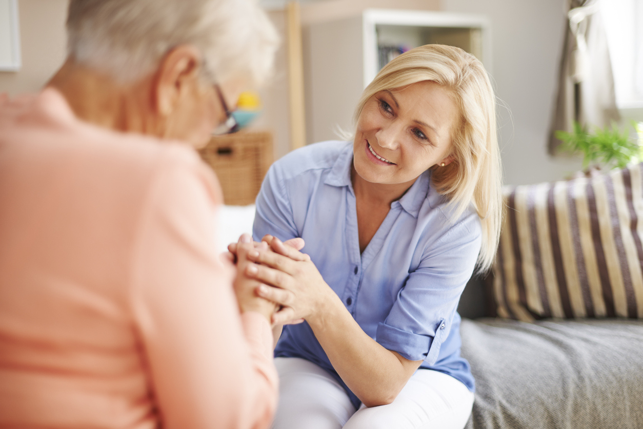 Woman in blue shirt talking and holding hands with an elderly woman indoors.