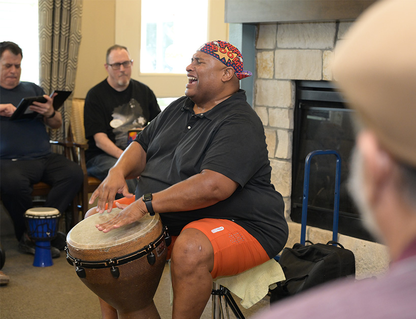 A man plays a djembe drum in a communal living space, engaging an audience.