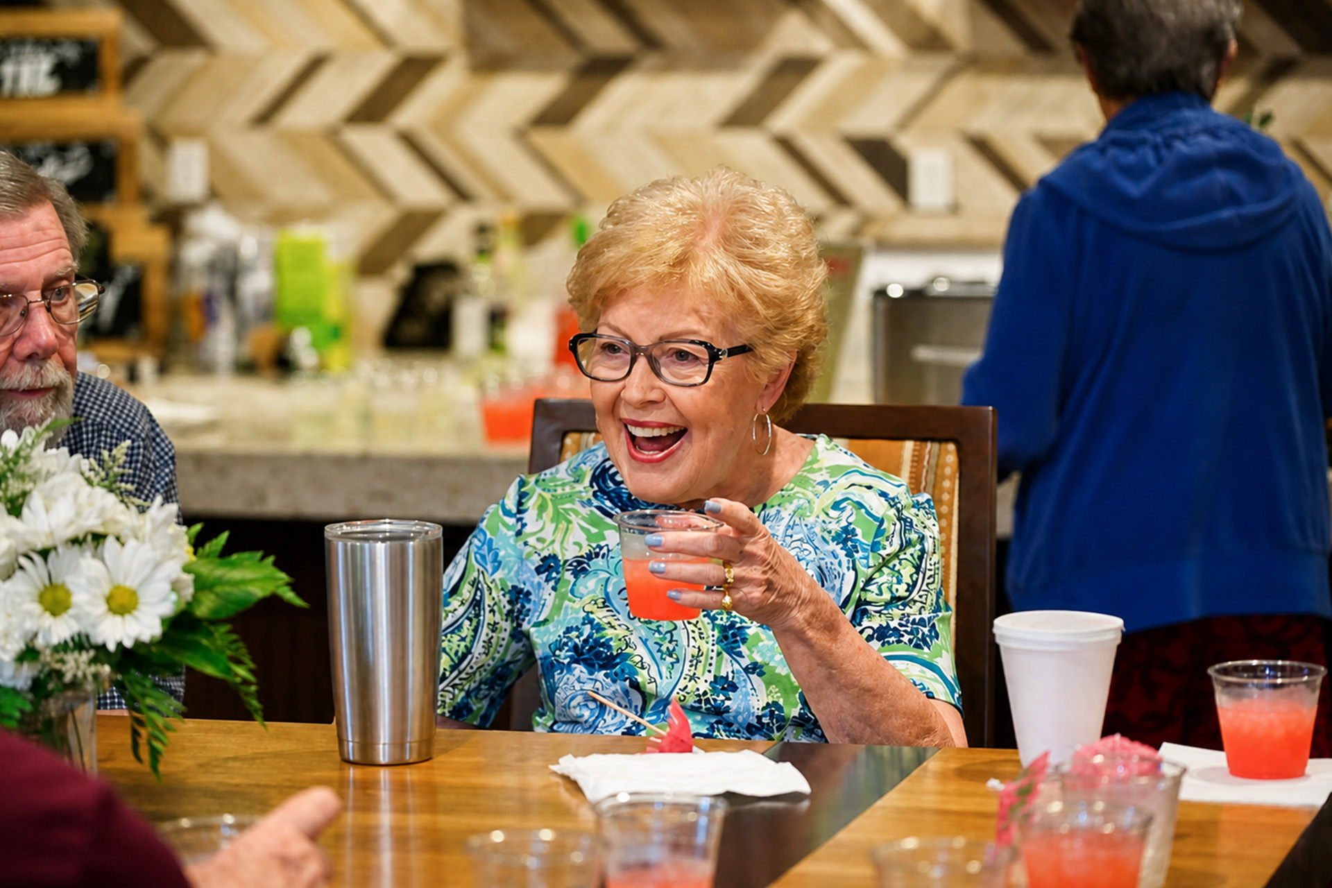 Senior woman at a table enjoying a drink with friends in a communal area.